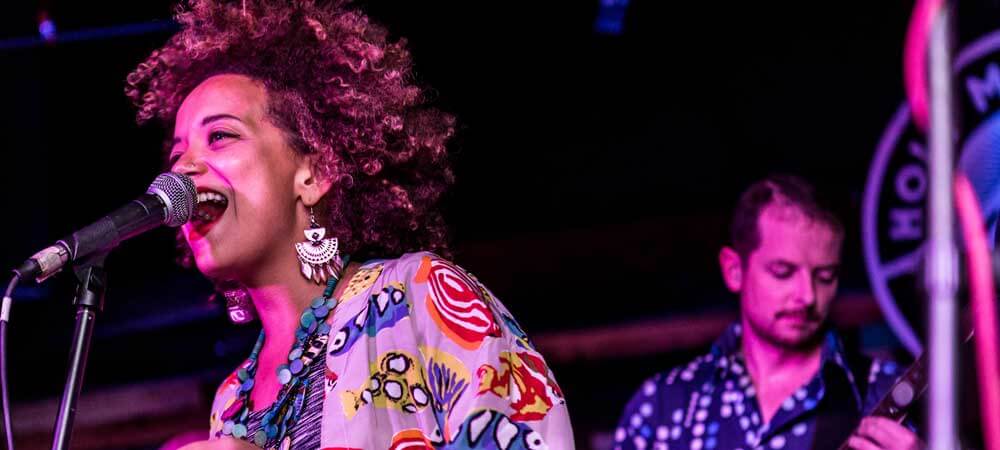 Photo of a woman singing on a stage with a man playing guitar in the background.