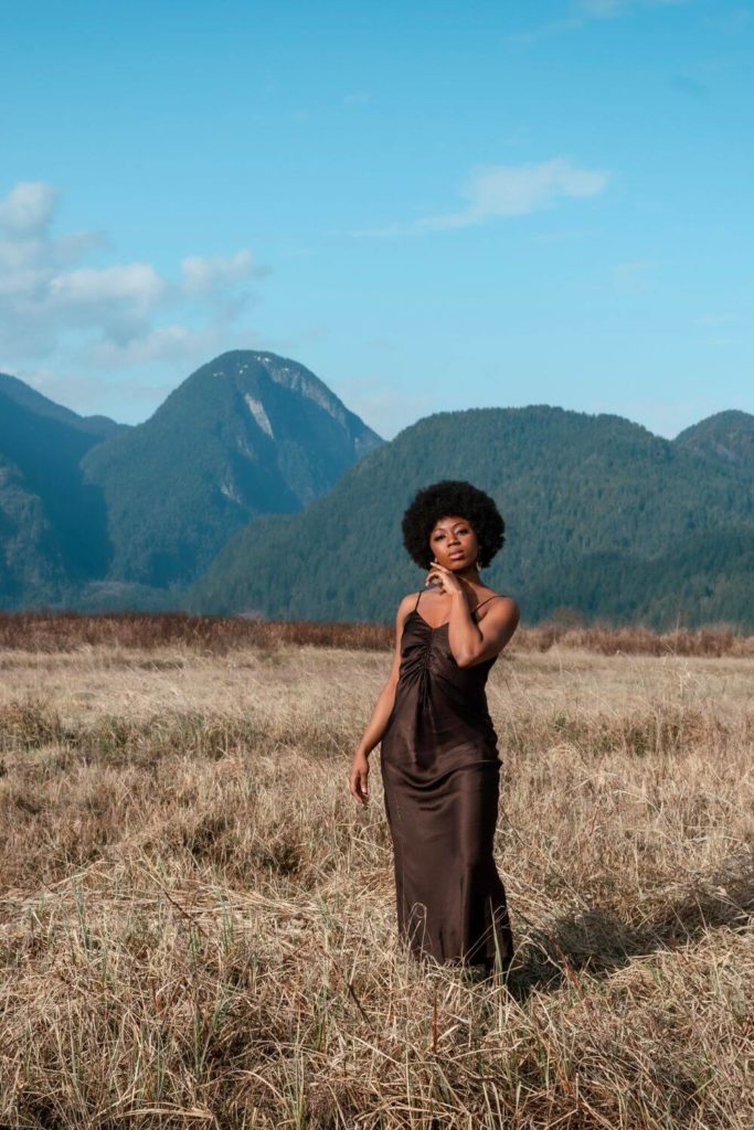 Photo of a woman wearing a chocolate brown silk slip dress, posing in a meadow with mountains in the background.