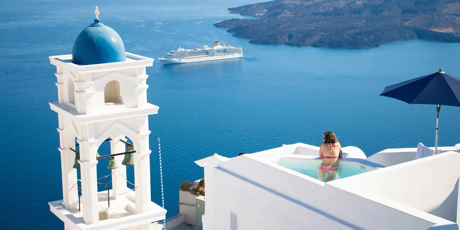 Ocean landscape view of Santorini, Greece showing a white bell tower with a blue dome, a woman in a rooftop pool, and a cruise ship in the distance.