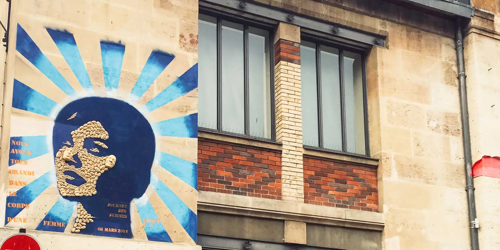 Street art on a building showing a silhouette of a woman's face made from smaller faces, with blue rays in the background.