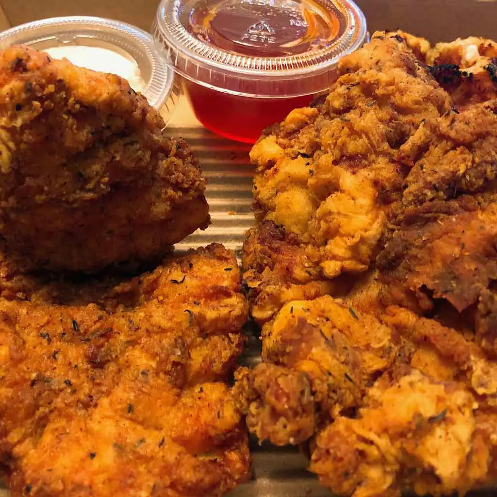 Closeup of a box of 2 pieces fried chicken and half a waffle, along with a cup of honey and a cup of butter.
