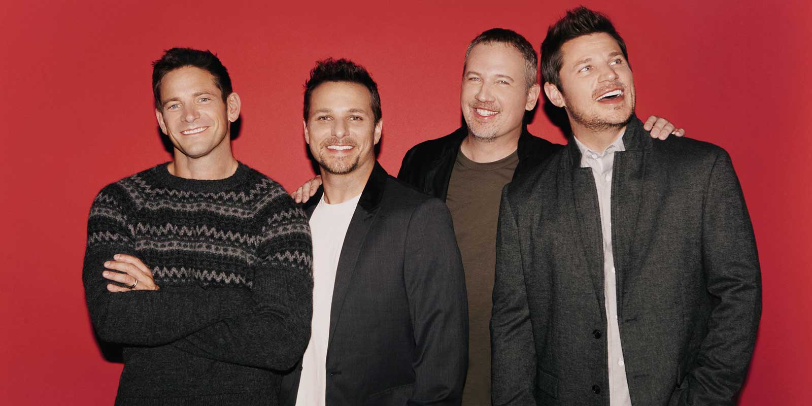 The 90s boys band 98 Degrees poses in front of a solid red background.
