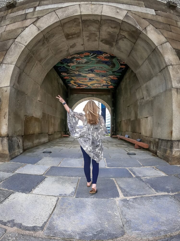 Travel blogger Meg Frost poses at Sungnyemun gate in Seoul, South Korea, flashing the peace sign.