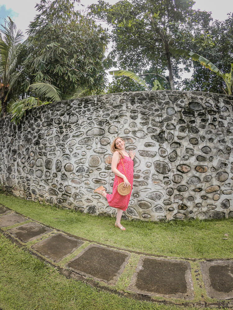 Travel blogger Meg Frost posing in front of a stone wall surrounded by tropical trees in Bali.