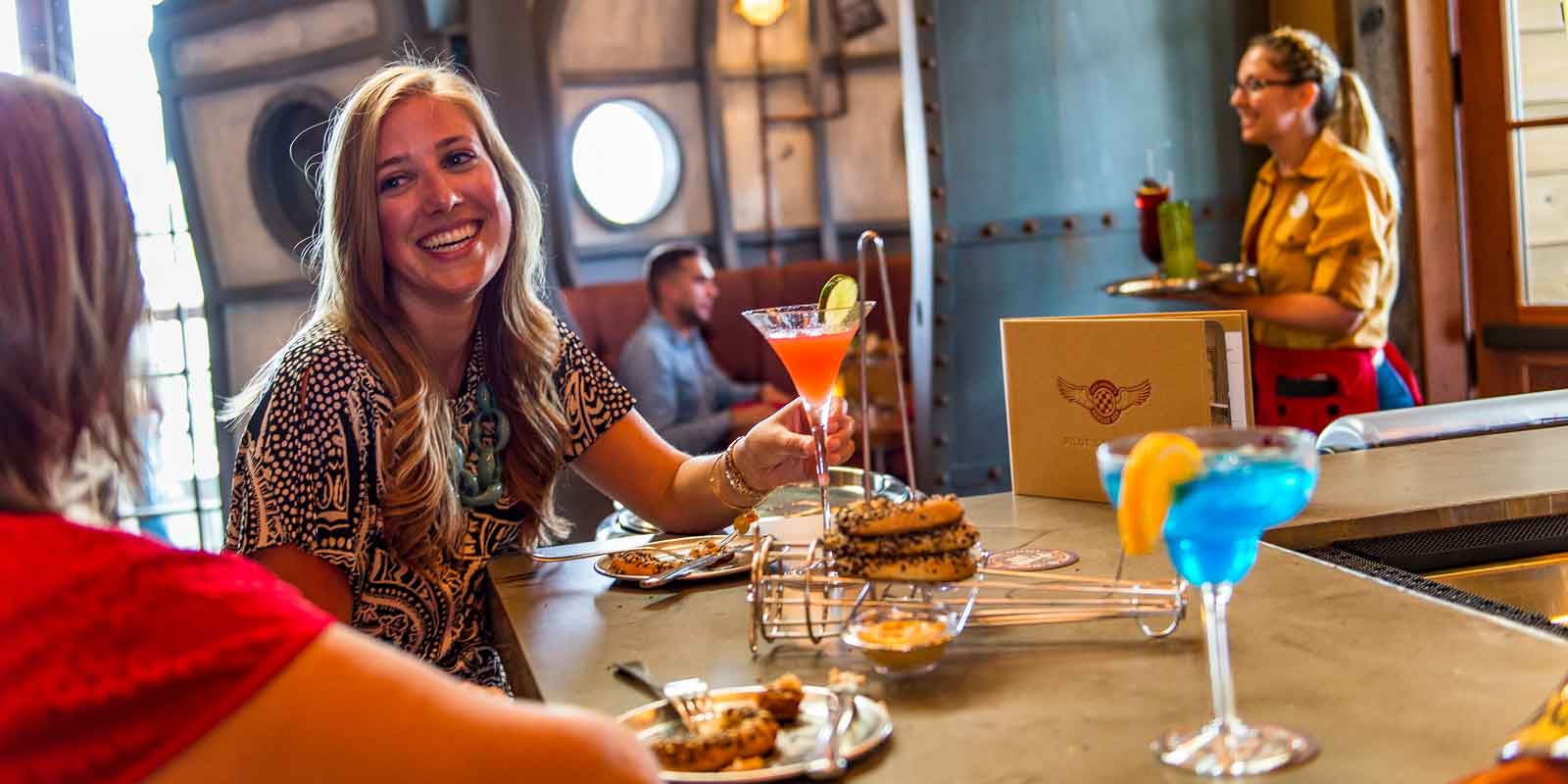 Close up of 2 women enjoying cocktails and an appetizer, with a cocktail waitress in the background, at Jock Lindsey's Hangar Bar.