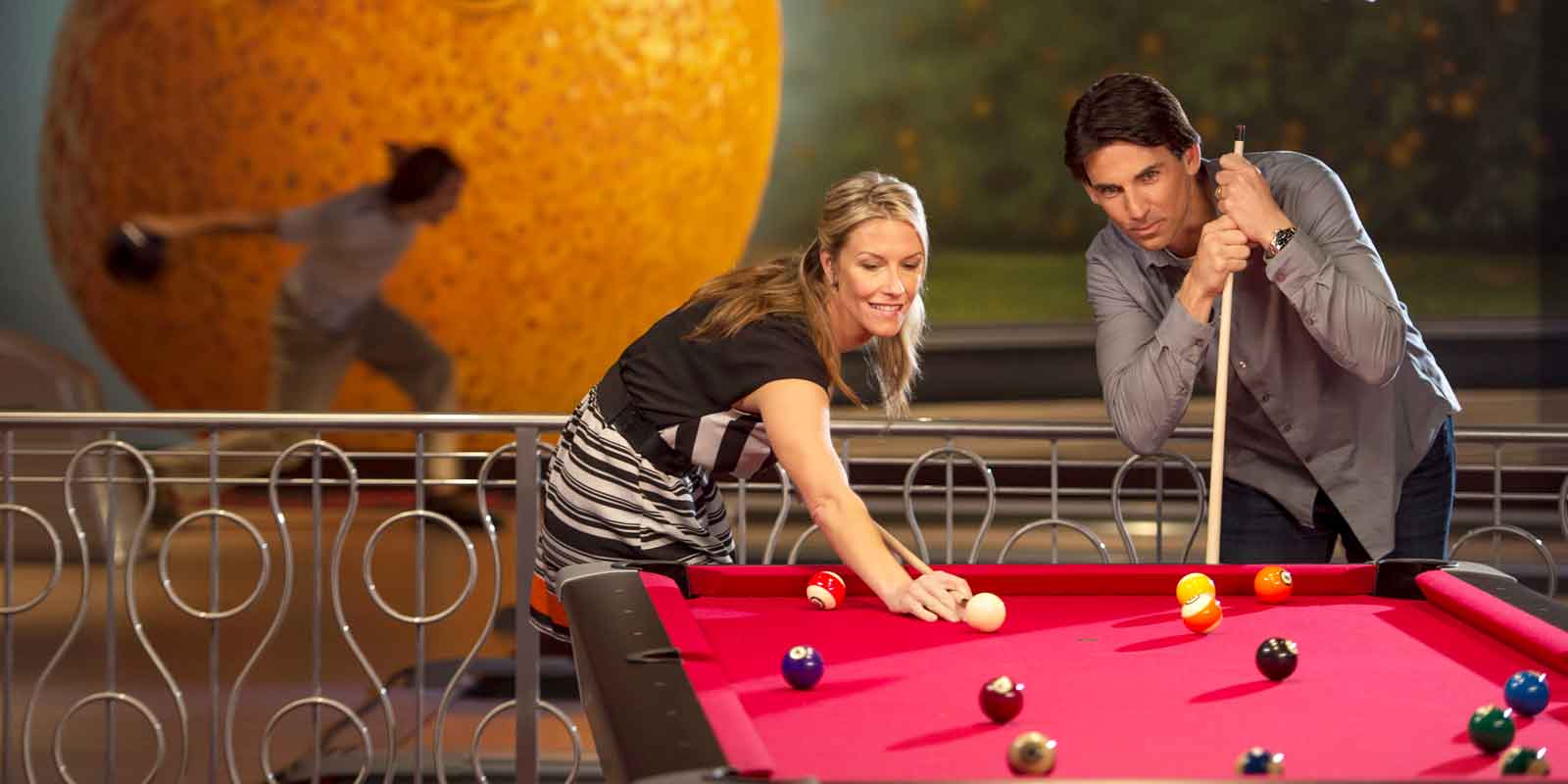 A woman and man play a game of pool on a red pool table, while a person in the background bowls.