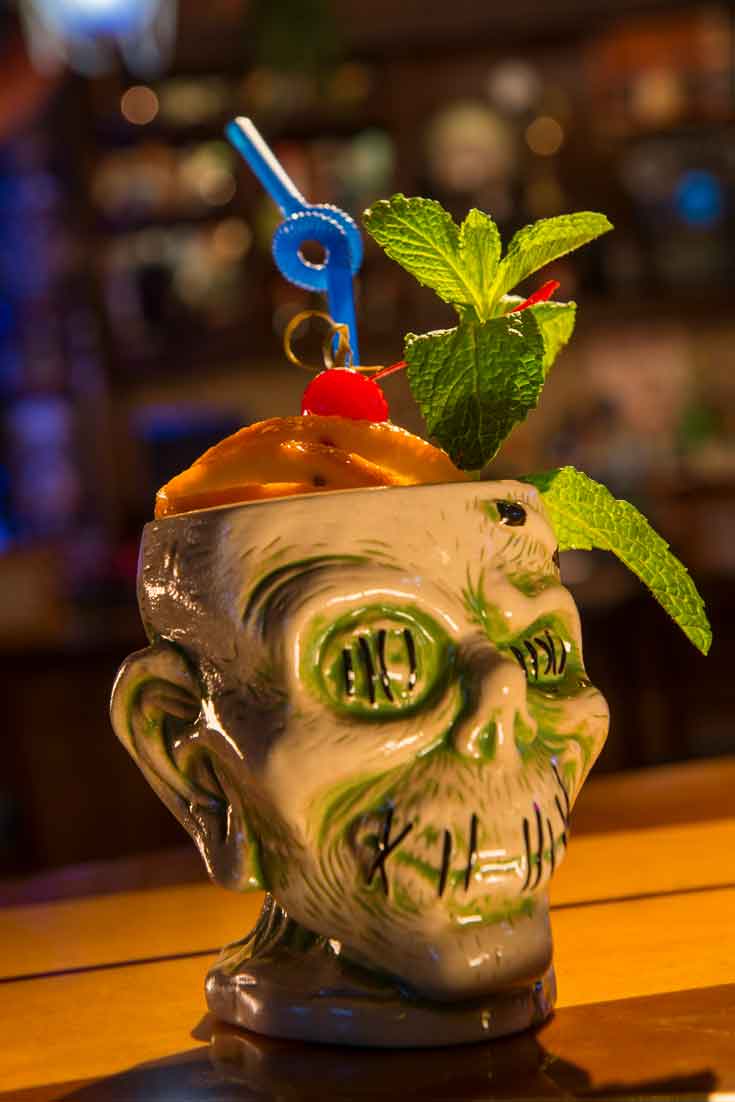 Closeup of a ceramic zombie cup filled with a cocktail and fruit, mint and a blue twisty straw.