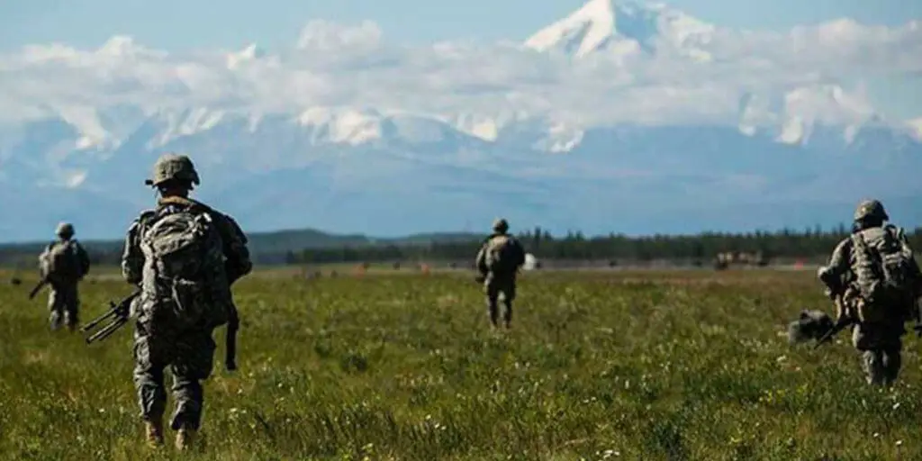 A group of US Army soldiers practice in a green field overlooking a mountain range.