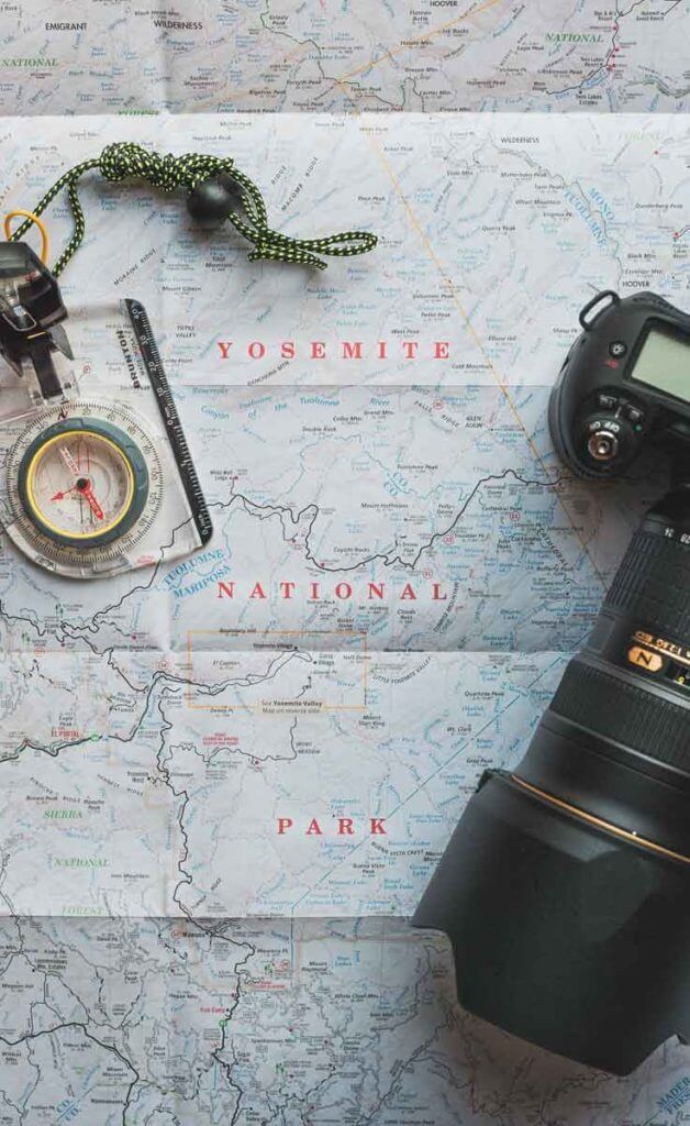 Flat lay photograph of a map of Yosemite National Park with a compass necklace and a dSLR camera with a long lens on top.