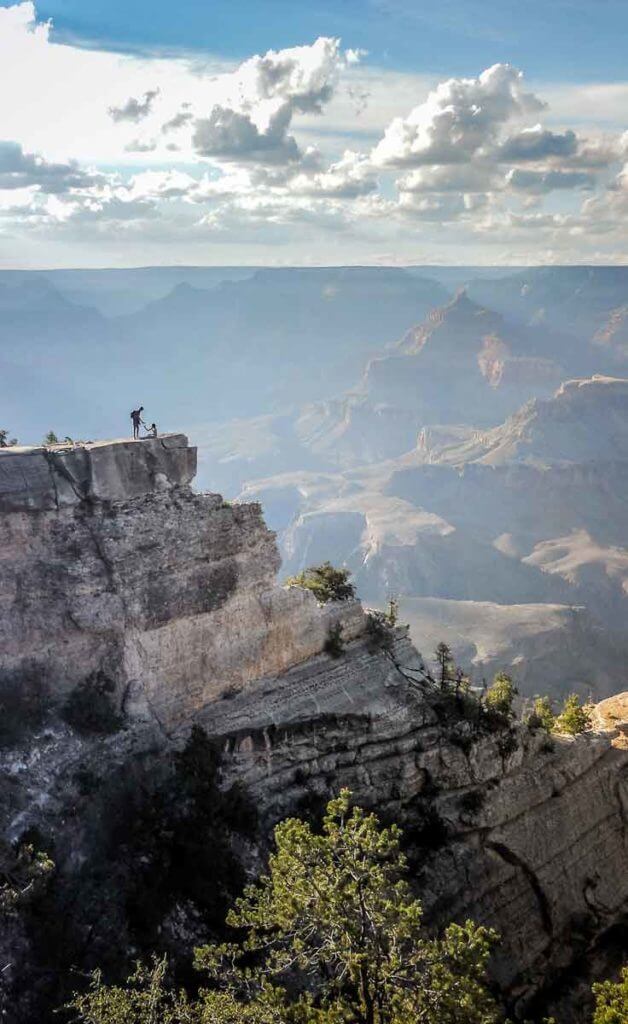 Landscape view of the Grand Canyon on a blue sky day, with a couple overlooking the canyon from a ledge in the distance.