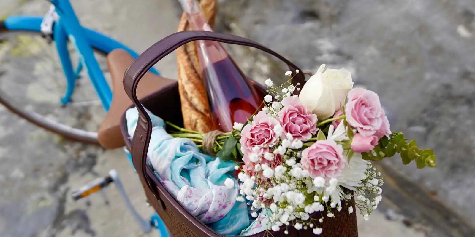 Close up of a basket on the back of a blue bicycle, filled with a baguette, bottle of pink wine, a blue and pink blanket or scarf, and a bouquet of pink carnations, white roses, and greenery.