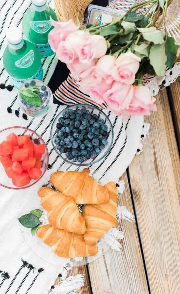 Flat lay photograph of a picnic spread, featuring a plate of croissants, a bowl of watermelon chunks, a bowl of blueberries, 2 bottles of San Pellegrino, a basket of pink roses, all on top of a white blanket with black stripes spread over a picnic table.