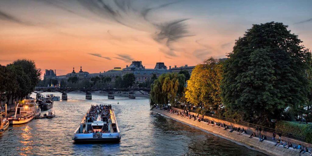 A Seine River cruise boat with passengers floats along the Seine River at sunset.