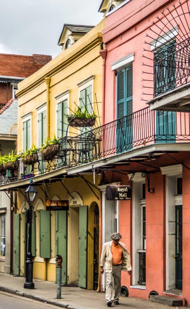 Photo of yellow and coral buildings in New Orleans' colorful French Quarter neighborhood, with an older gentleman in a tan suit and orange shirt walking by.