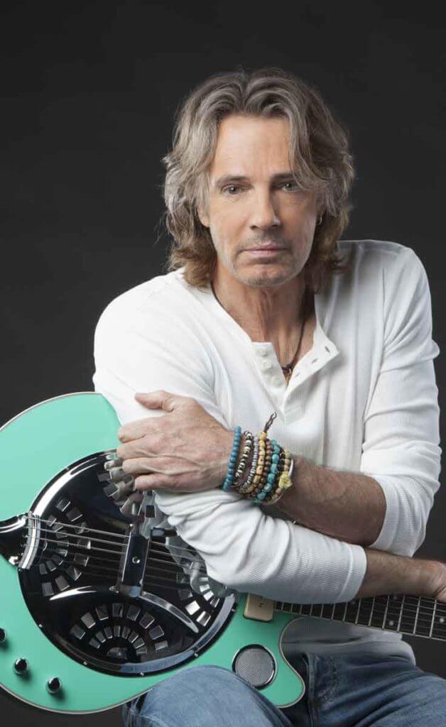 Photograph of musician Rick Springfield wearing a white long sleeve henley tee and jeans, leaning over an aqua guitar.