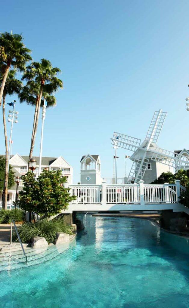 Photograph of a section of the Stormalong Bay pool at Disney World, featuring a white bridge, turquoise water, a windmill, and part of a resort building in the background.