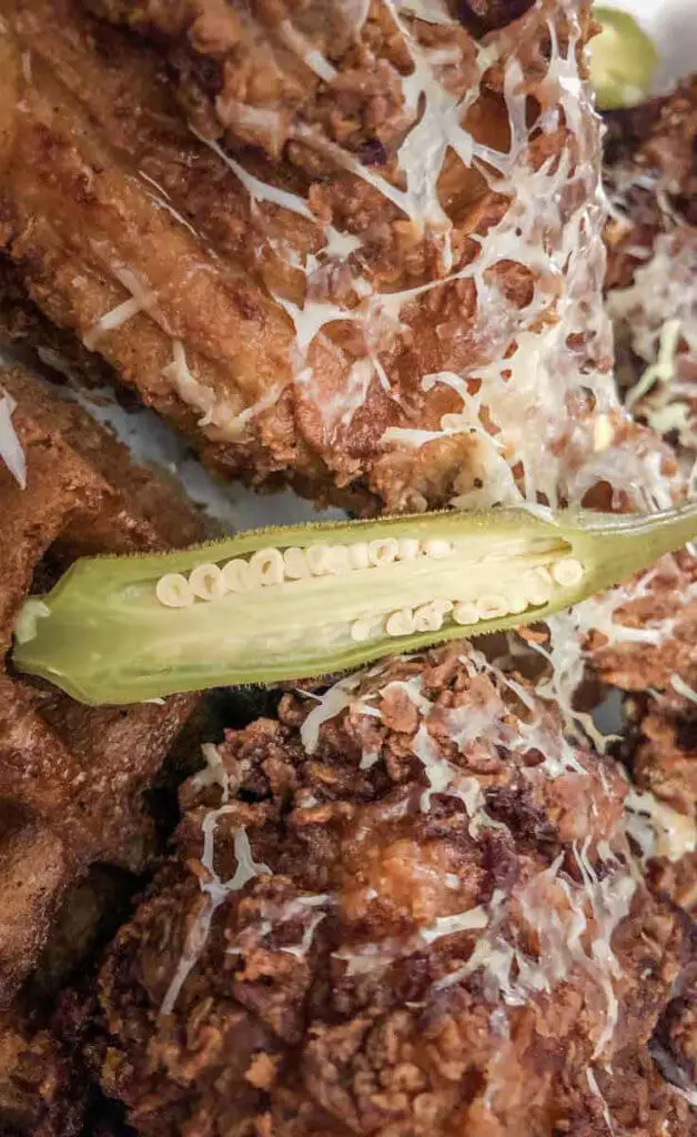 Closeup shot of fried chicken covered in shredded cheese and okra, on top of waffles.