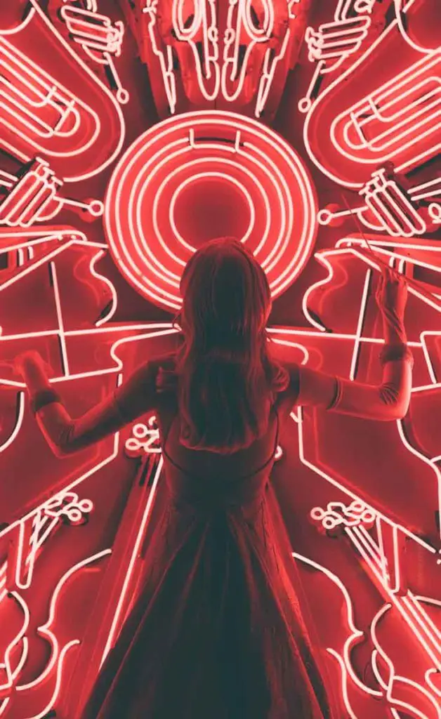 Woman in a dress examines a red neon sign that features Jazz instruments, like a piano and trumpet.