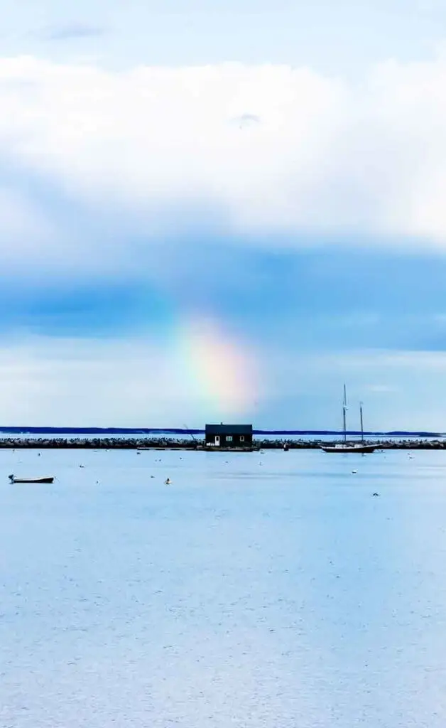 Landscape view of the ocean in Provincetown and a rocky patch of land with a small shack. The sky is bright blue with a rainbow stretching out of a cloud above the shack.