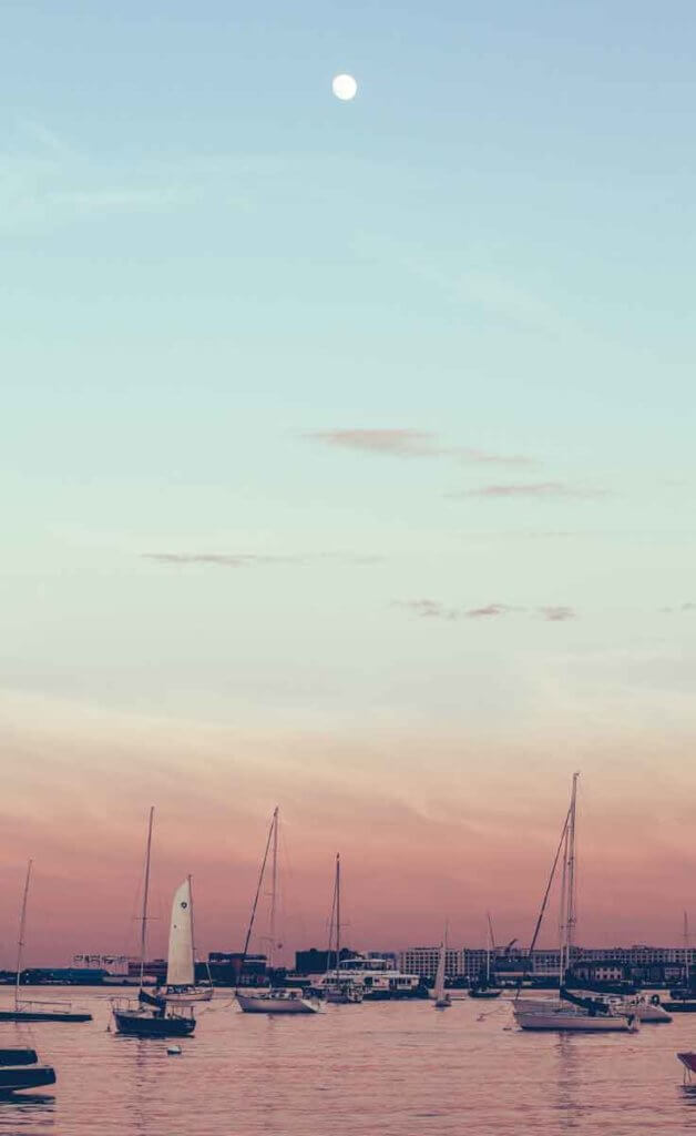 Sailboats along the Boston Harbor with the city of Boston in the background. The sky is light blue, fading to pinkish orange.