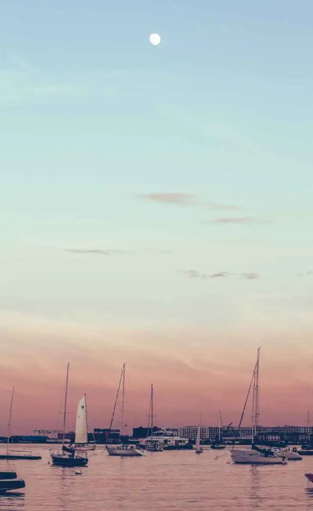 Sailboats along the Boston Harbor with the city of Boston in the background. The sky is light blue, fading to pinkish orange.