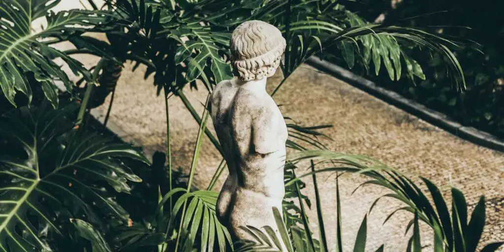 Armless statue amidst large leaves and plants with a tan stone sidewalk nearby.