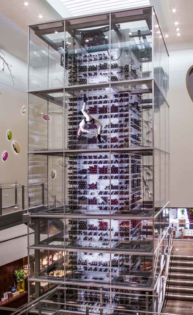 Photo of the massive tower of wine enclosed by glass at Aureole at Mandalay Bay. A woman is suspended from the top in a harness in an acrobatic pose.