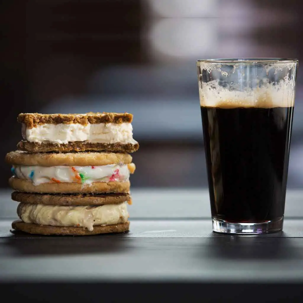 A stack of 3 ice cream cookie sandwiches sits next to a pint of dark beer.