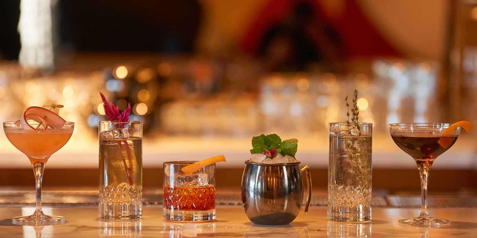 Closeup of a bar featuring 6 cocktails, all with botanical sprigs or other fun garnishes.