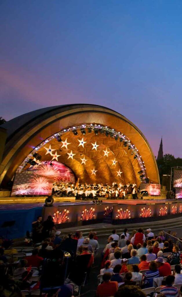 Boston Pops Orchestra performing on July 4th in the Hatch Shell stage along the Charles River in Boston at dusk.