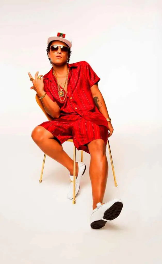 Promo photograph of R&B singer Bruno Mars. Mars is wearing a red silk button up top and shorts with lots of gold jewelry, white loafers, and a white baseball cap.