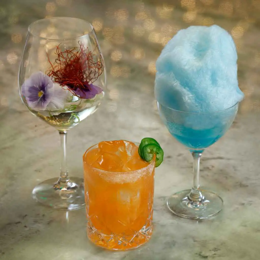 Closeup of 3 cocktails, one in a deep wineglass with a violet garnish, another bright orange cocktail with jalapeño garnish, and the last with a tall puff of baby aqua cotton candy.
