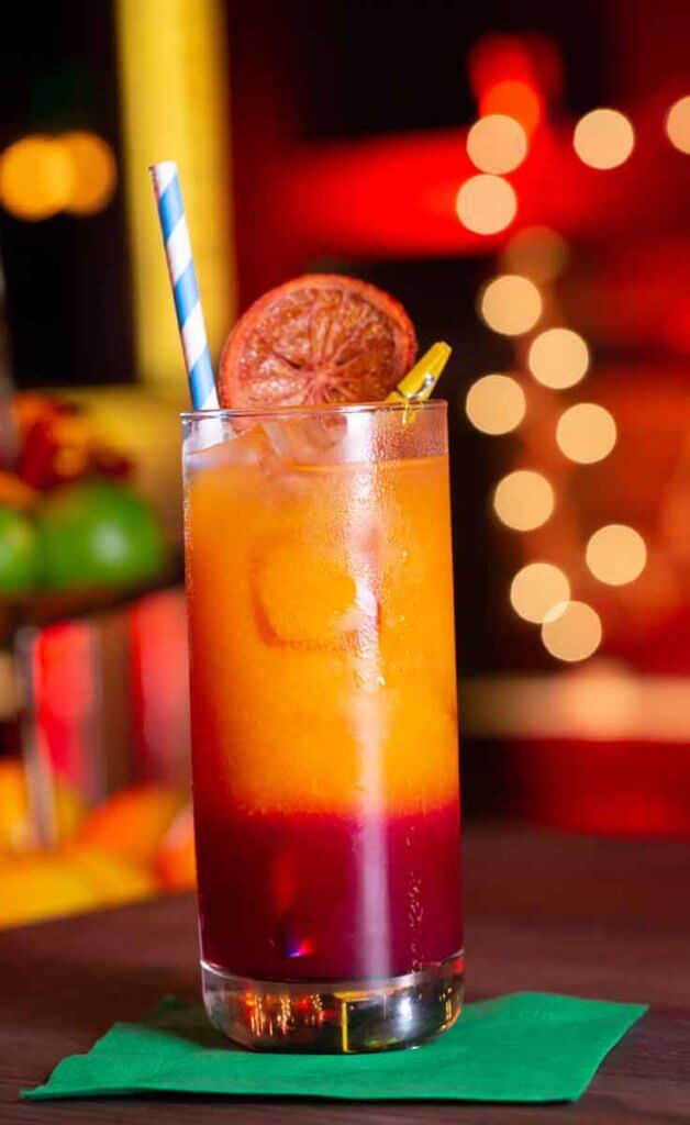 Closeup of a highball glass filled with a cocktail that is cherry red at the bottom and orange at the top, garnished with a dried blood orange and a blue and white striped paper straw.