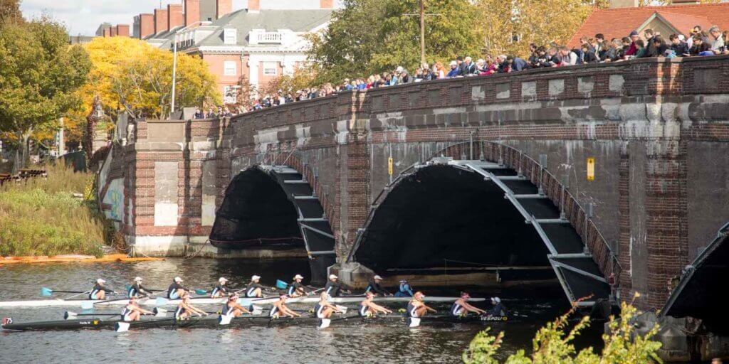 2 8-person rowing boats glide through the Charles River, while crowds watch from a bridge above.