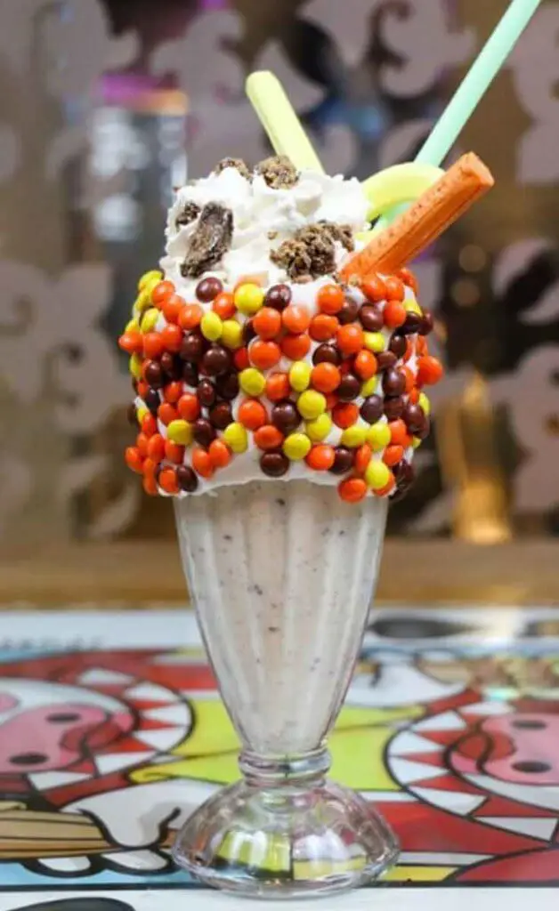 Closeup of a booze-filled milkeshake with Reese's pieces candies surrounding the rim of the glass and a tall mountain of whipped cream.