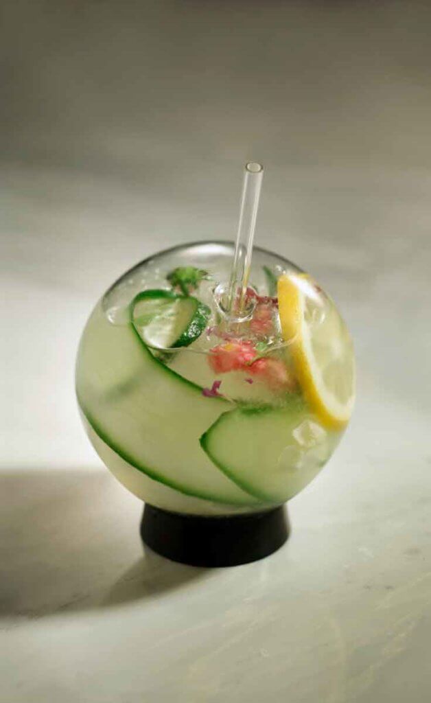 Closeup of a spherical glass filled with a gin cocktail, long strips of cucumber, lemon slices, and a flower garnish.