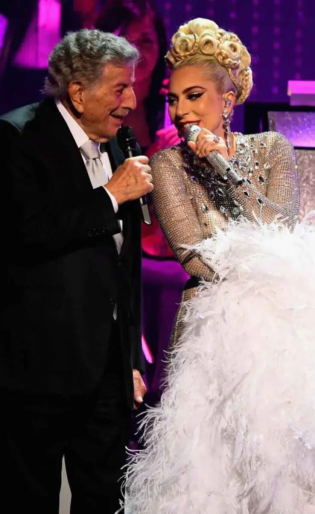 Singers Tony Bennett, in a black suit, and Lady Gaga, in a silver sparkly and feather adorned gown, sing on stage at the MGM Park Theater.