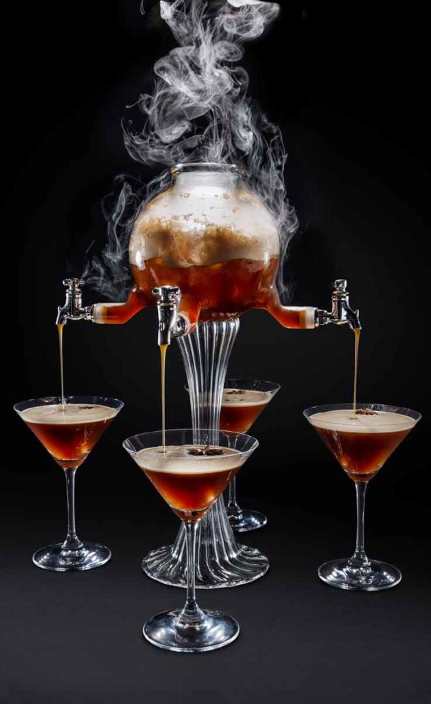 Photograph of a steaming and bubbling drink dispenser with 4 spouts, all pouring an espresso cocktail into martini glasses.