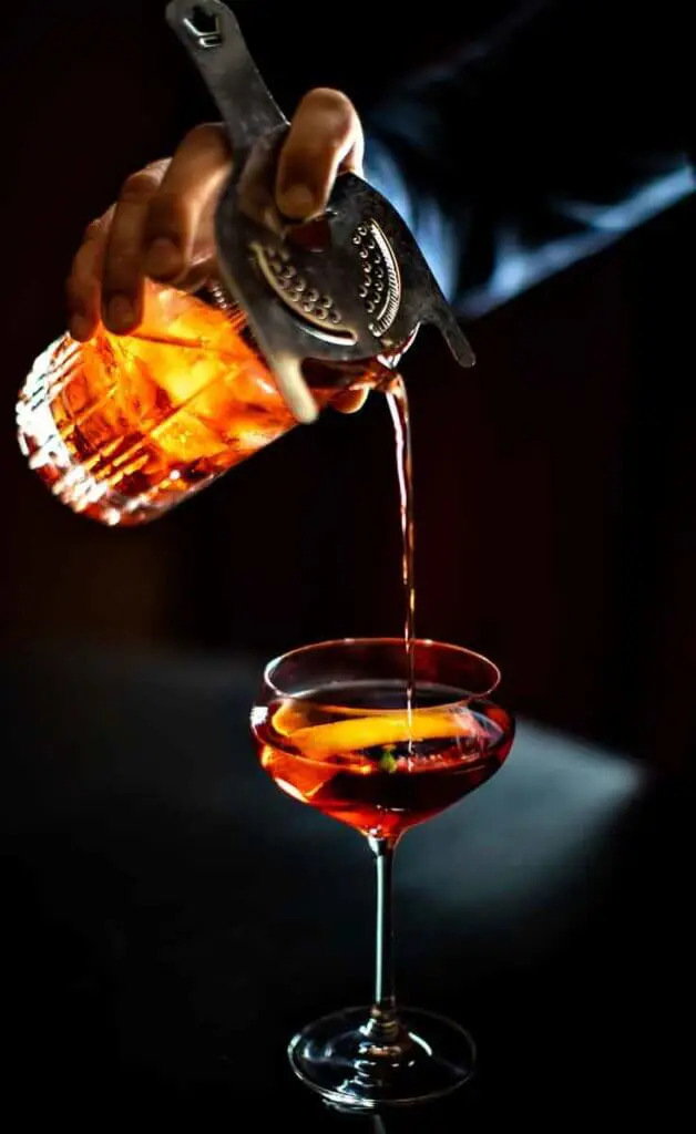 Closeup of a person pouring a whisky-based cocktail through a glass with a strainer into a tall stemmed cocktail glass.