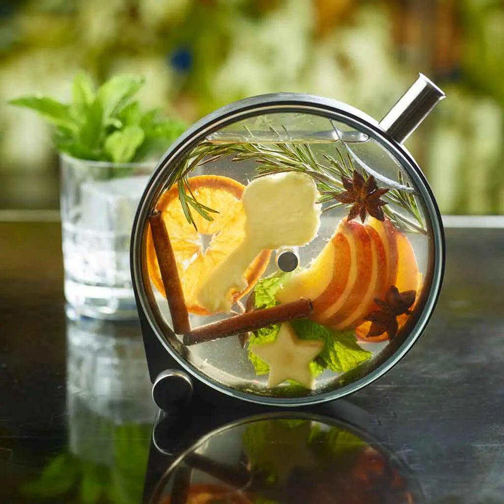 Closeup of a cocktail in a round, cask-link container. Inside are cinnamon sticks, a sprig of rosemary, orange slices, apple slices, and other garnishes.