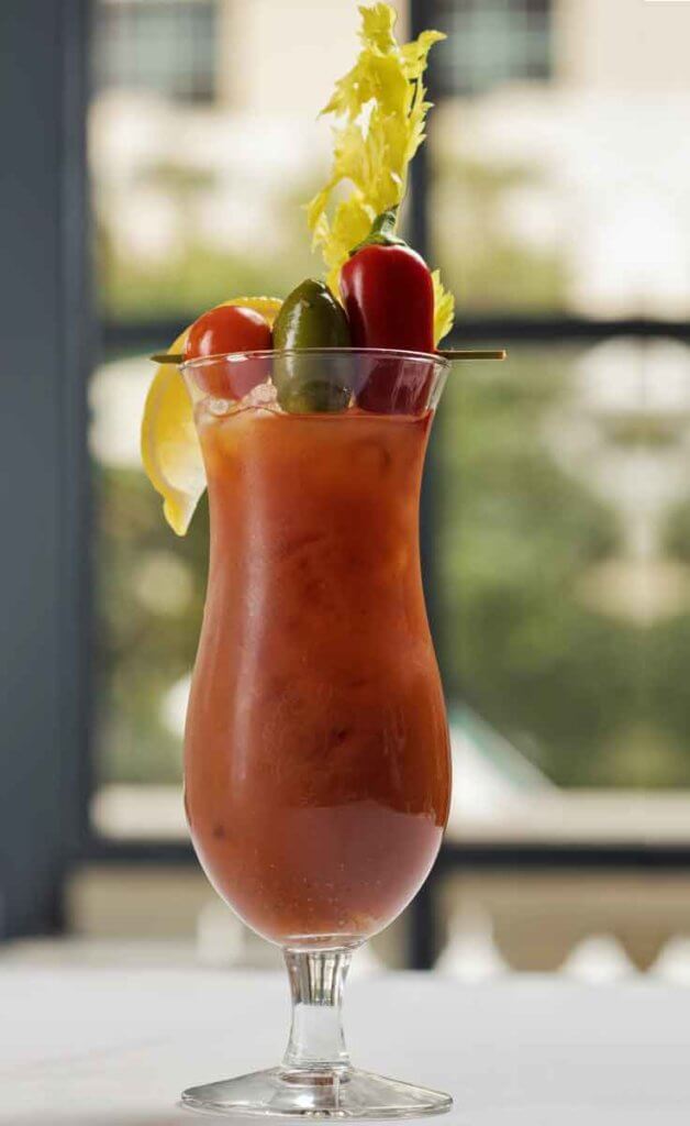 Closeup of a tall hurricane glass with a Bloody Mary cocktail, complete with a lemon wedge, stalk of celery, and a skewer with a tomato, olive, and chile pepper.