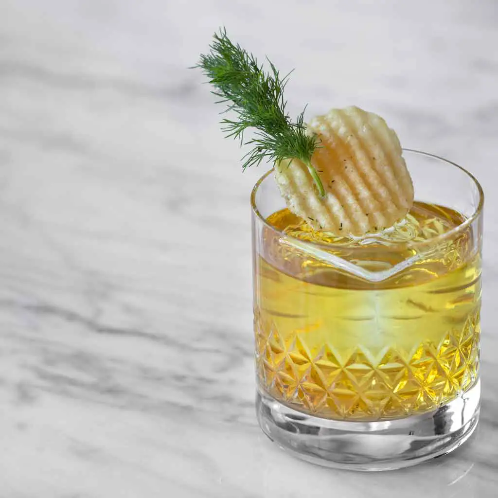 Closeup of a short glass with a yellow tinted cocktail garnished with a rippled potato chip and sprig of dill.
