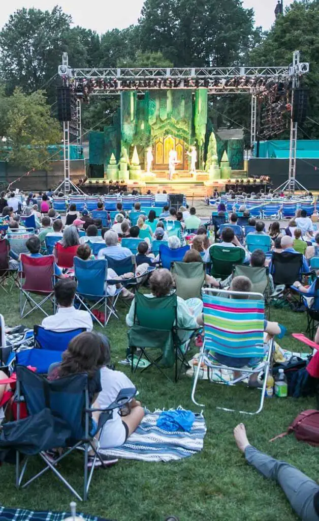 People in lawn chairs on the Boston Common watch actors on an outdoor stage.