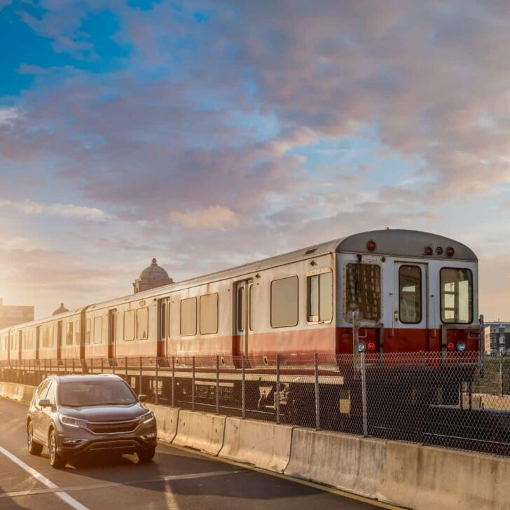 Photo of a Red line train crossing a bridge over the Charles River between Boston and Cambridge, MA with a car driving next to it.