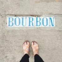 Photograph pointing down to the ground, showing a woman's feet in sandals and tiles embedded in a sidewalk that spell out 