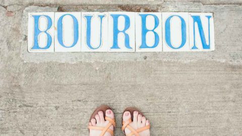 Photograph pointing down to the ground, showing a woman's feet in sandals and tiles embedded in a sidewalk that spell out "Bourbon."