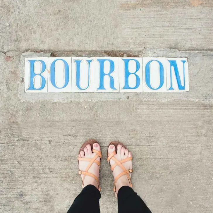 Photograph pointing down to the ground, showing a woman's feet in sandals and tiles embedded in a sidewalk that spell out "Bourbon."