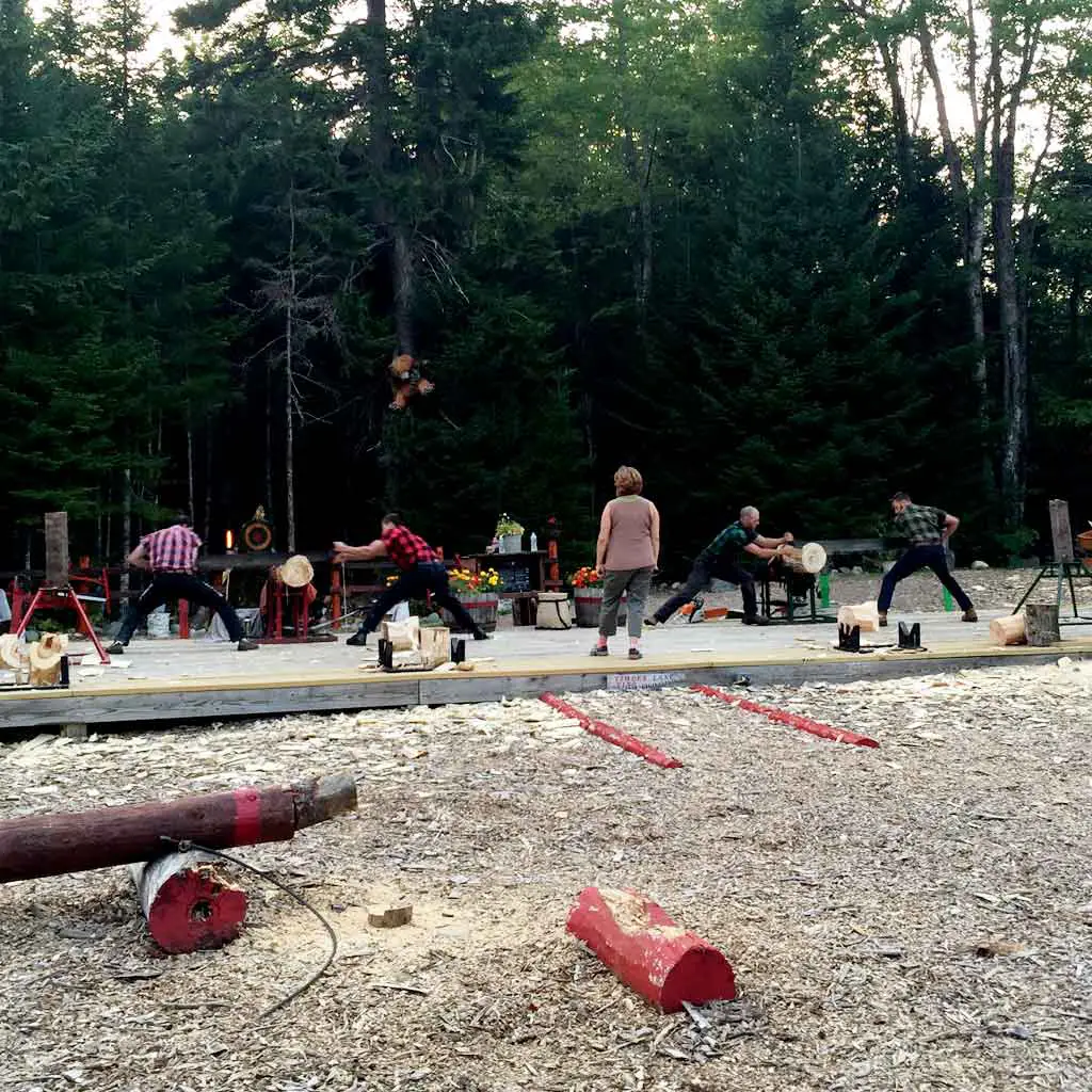 Two teams of 2 men competing in sawing a log while a woman overlooks the contest.