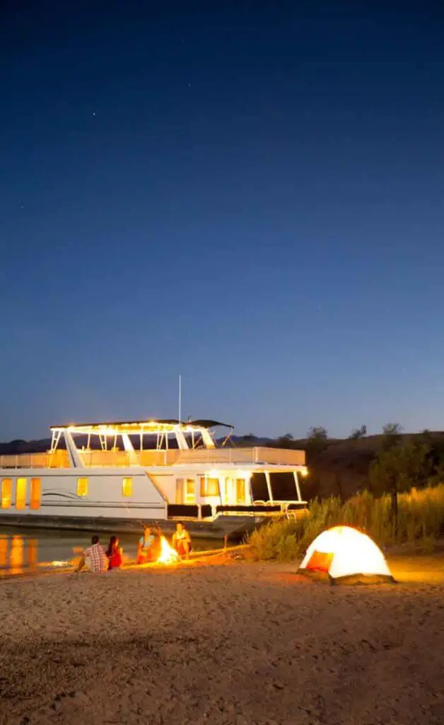 A boat docked along Lake Havasu at night with people sitting around a bonfire and a tent on the shore.
