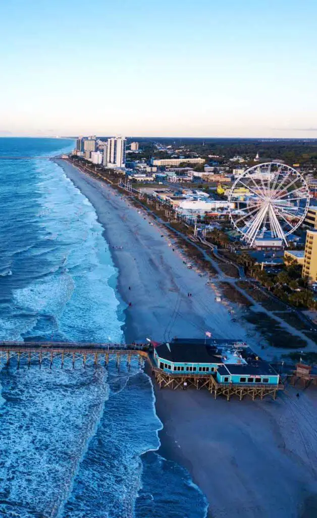 Aerial view of a long stretch of beach with a pier stretching into the ocean, tall resort buildings, and a ferris wheel dotting the shore.